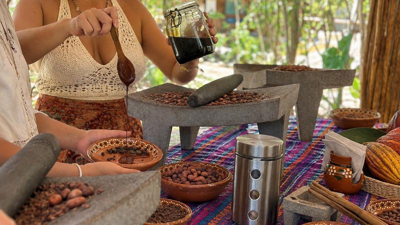 Guests grinding cacao on stone metates and pouring fresh chocolate over the nibs at Tierra Maya, Cozumel, with whole cacao pods on the table